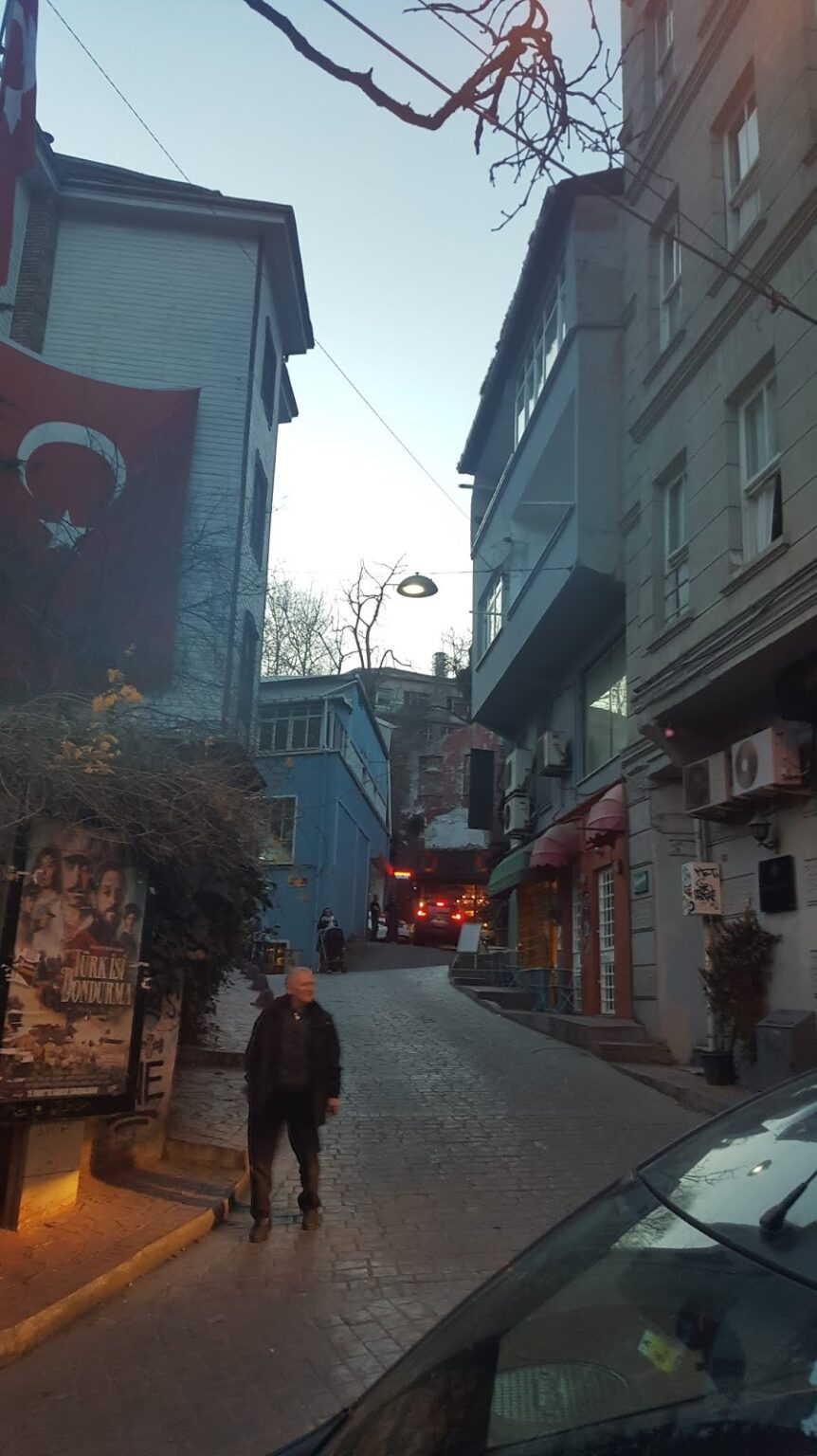 Cihangir semtinde dar bir sokak, binaların arasında bir adam yürüyerek ilerliyor. Arkada Türk bayrağı ve çeşitli dükkanlar görünmekte.