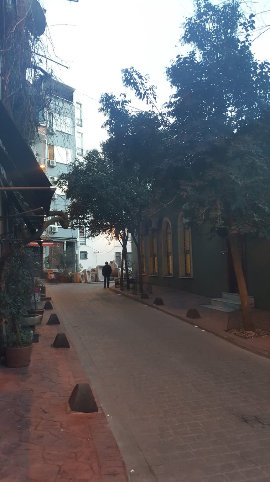 Cihangir semtinde ağaçlarla çevrili dar bir sokak, arka planda binalar ve sezgisel aydınlatmalar gözüküyor.
