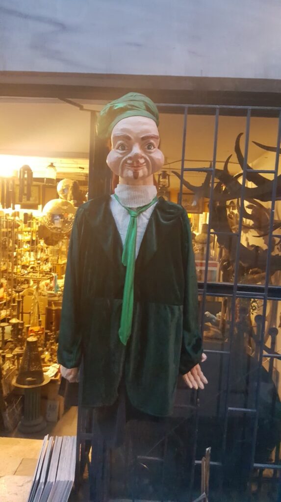 Bir dükkanın önünde, yeşil bir kıyafet giymiş ve başında bir şapka bulunan büyük bir kukla.