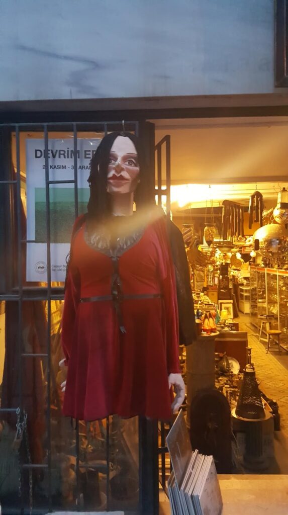 Bir dükkanın önünde asılı, uzun siyah saçlı ve kırmızı bir elbise giymiş bir manken. Arkada ilginç objelerle dolu bir antikacı gözüküyor.