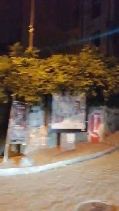 Gece çekilmiş bir sokak görüntüsü; arka planda ağaçlarla kaplı bir alan ve çeşitli afişlerin bulunduğu bir duvar.