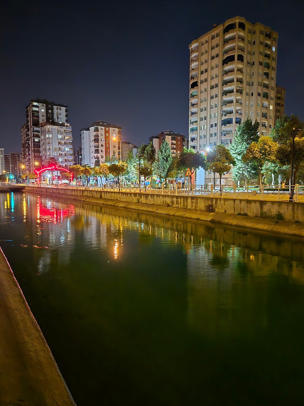 Gece çekimi, aydınlatılmış bir nehir kenarı ve çevresindeki apartman binaları.