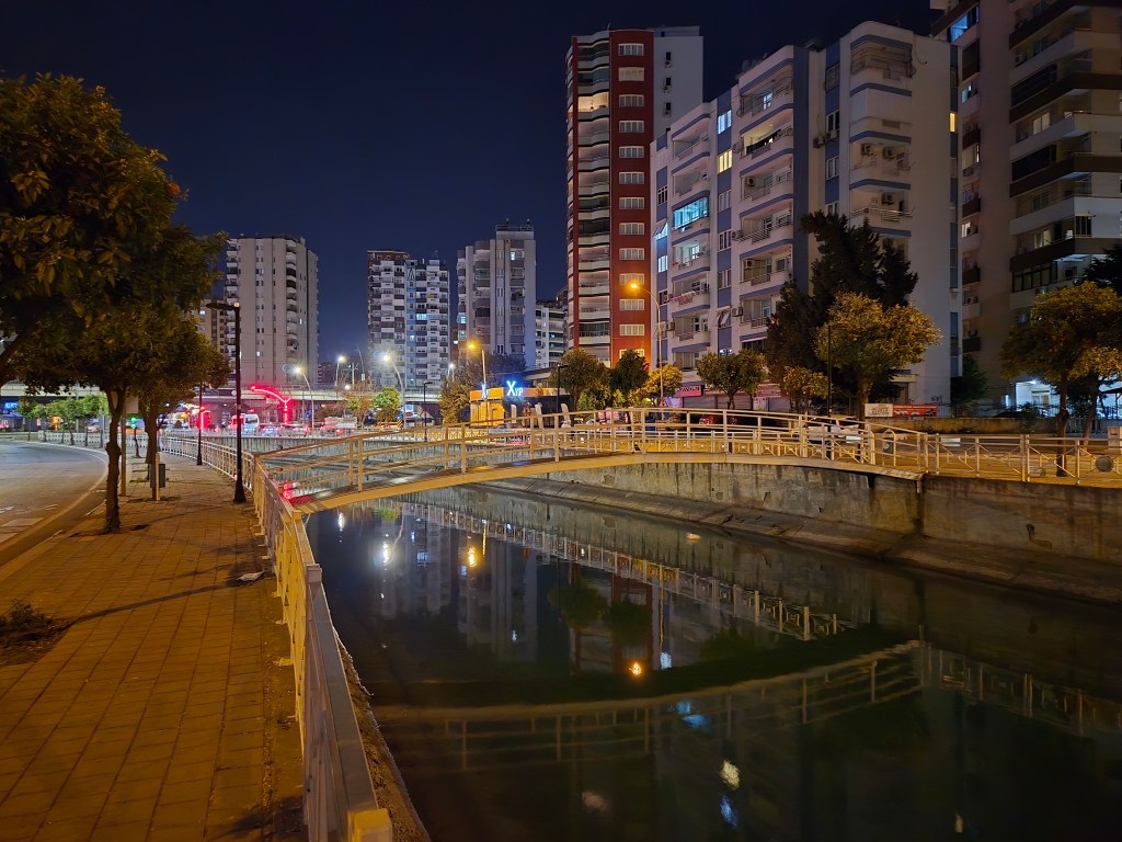 Gece çekimi, bir şehir sokak manzarasında su kenarındaki yaya köprüsü, çevredeki modern binalar ve ağaçlar görünmektedir.