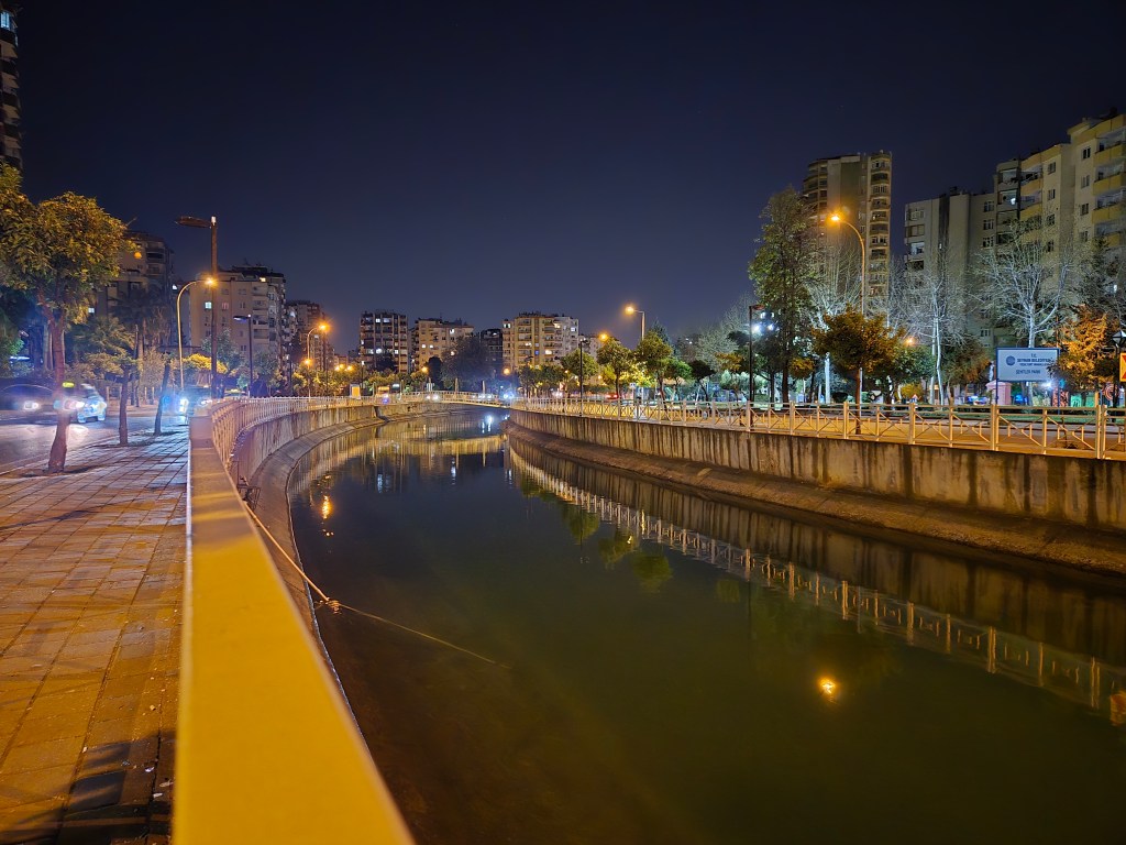 Gece çekimleri ile aydınlatılmış bir şehir manzarası; su kenarında yürüyüş yolu, binalar ve sokak lambaları.