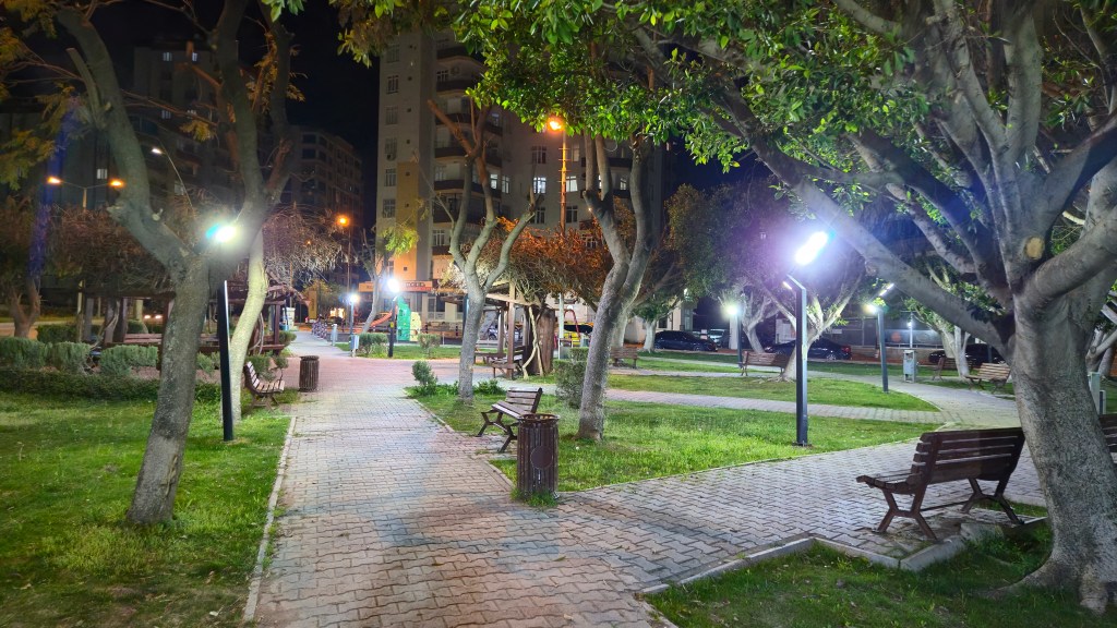Gece çekiminde bir park görünümü, ağaçlar ve bankların gölgesi ile birlikte sokak lambalarının aydınlattığı yollar.