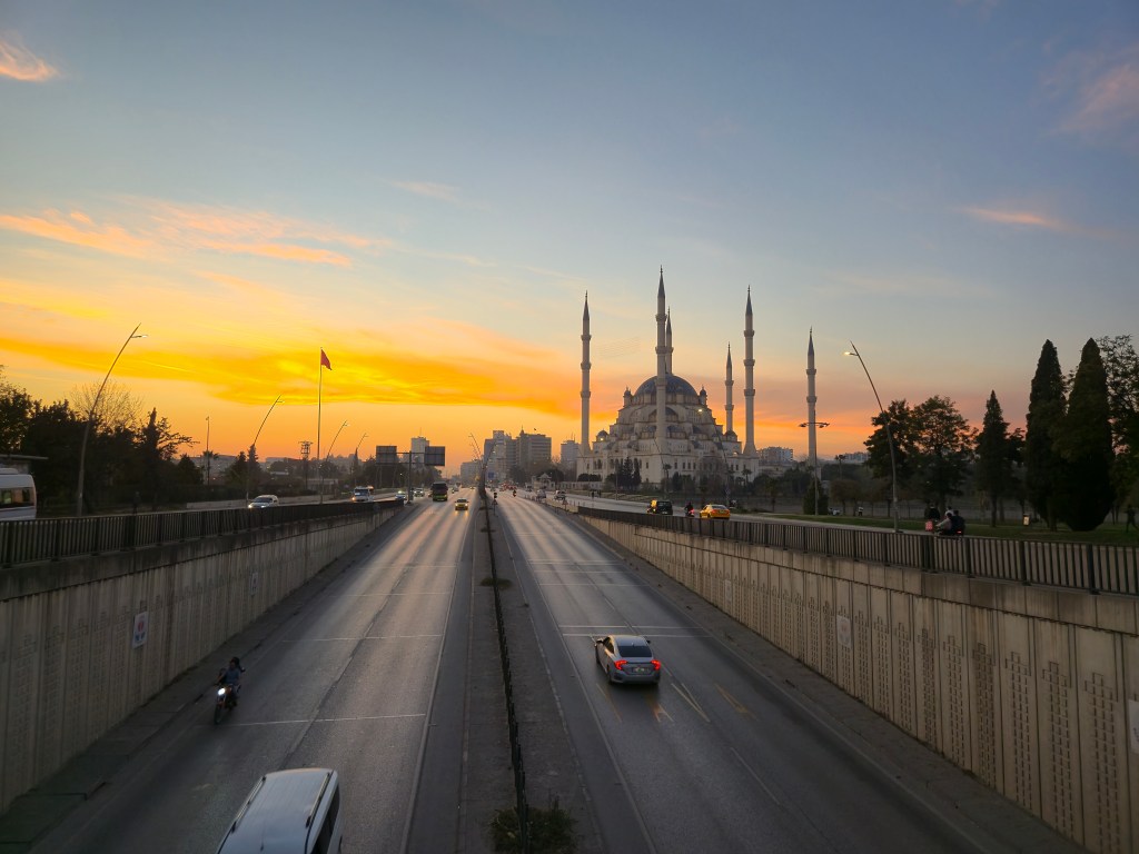 Güneşin batışıyla aydınlanan şehir manzarasında, ön planda bir yol ile arka planda bir cami yer almakta. Gökyüzü turuncu ve mavi tonlarında renklerle dolu.