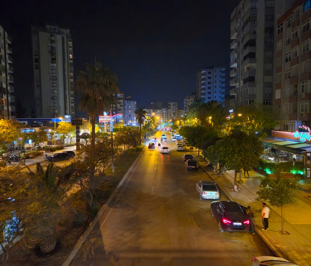 Gece çekimi, aydınlatılmış sokak ve binalar arasında ilerleyen araçlar ile palmiyeler