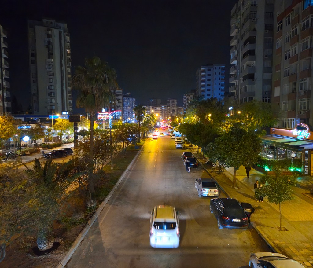 Gece çekiminde aydınlatılmış bir cadde, ağaçlar ve binalarla çevrili, birkaç araç yolda ilerliyor.