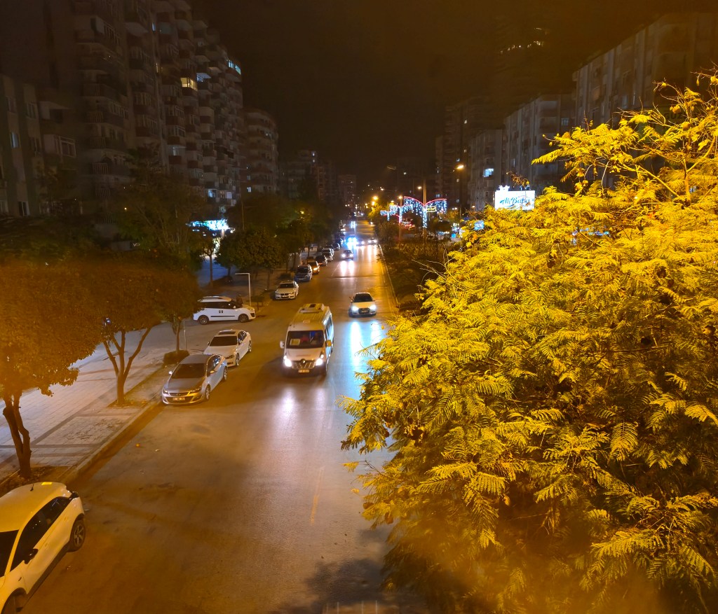Gece çekimi, caddede ilerleyen araçlar ve aydınlatılmış binalar ile dolu bir manzara.