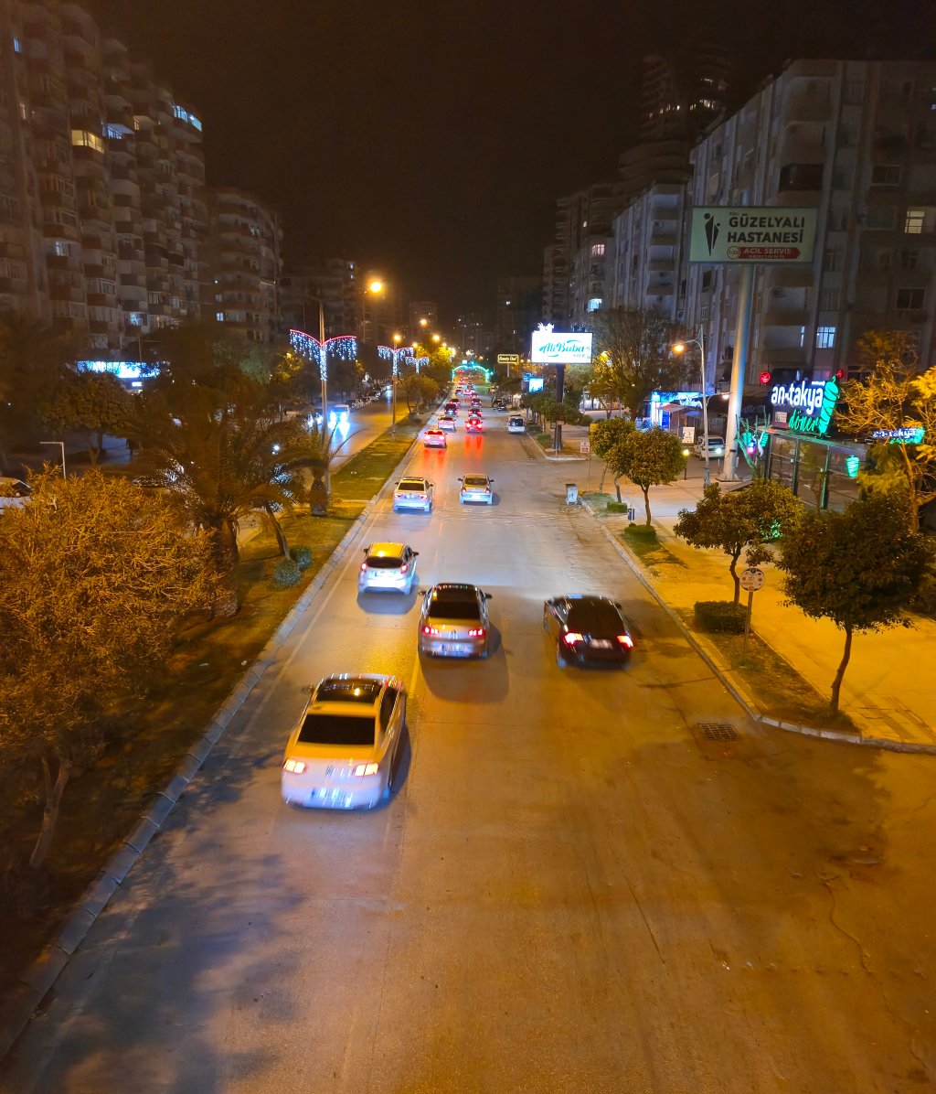 Gece görülen bir cadde, çeşitli araçların hareket ettiği aydınlatmalı bir ortam.