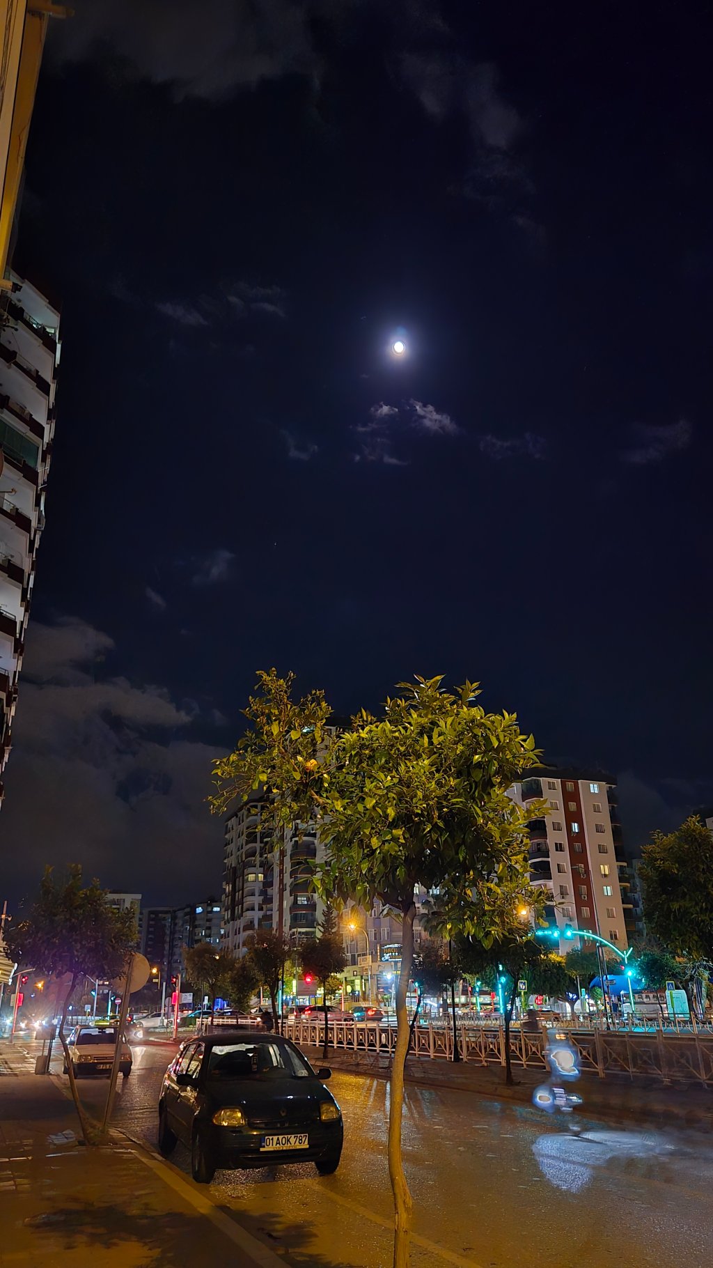 Gece görüntüsünde ay, bulutlar ve aydınlatılmış bir sokak manzarası, park etmiş bir araba ve çevresindeki binalar görünmektedir.