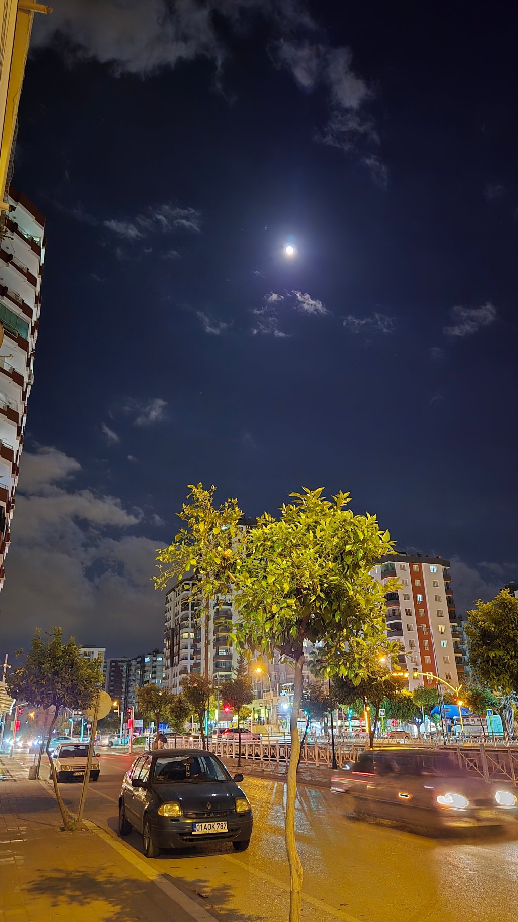 Gece çekimi; aydınlatılmış bir cadde, park halindeki bir otomobil ve ağaçlar. Arka planda apartmanlar ve sokak lambaları görünmekte.