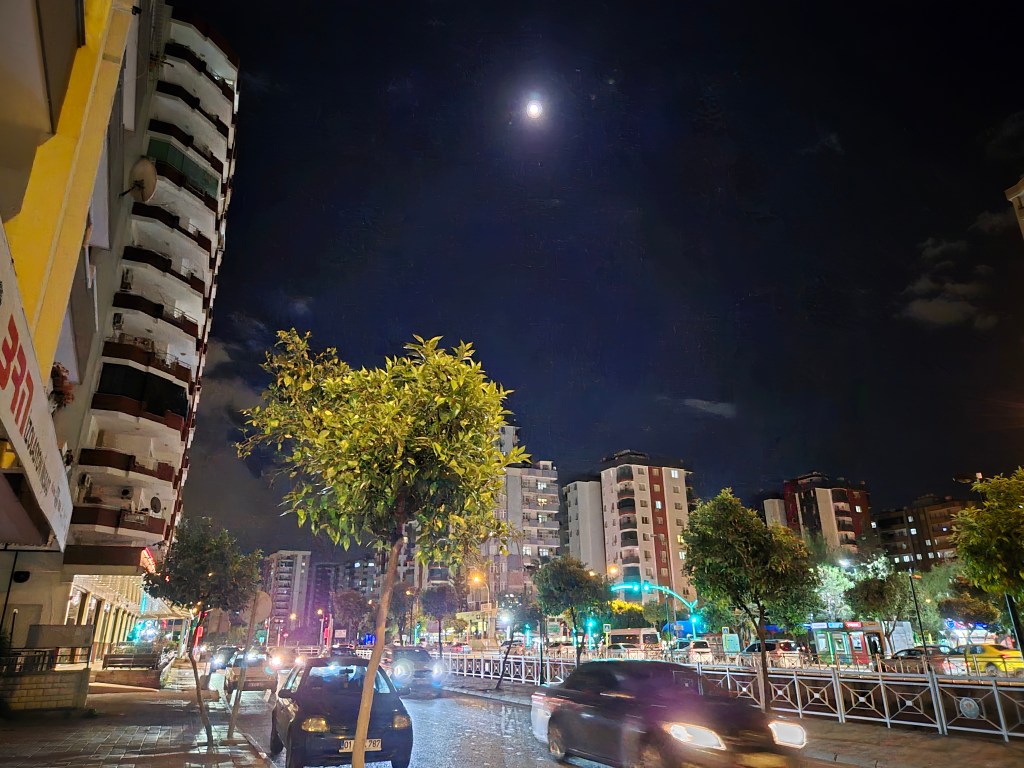 Gece manzarası, ay ışığı altında aydınlatılmış bir şehir sokak görüntüsü. Binalar, ağaçlar ve aracın geçişiyle hareketli bir atmosfer yaratıyor.