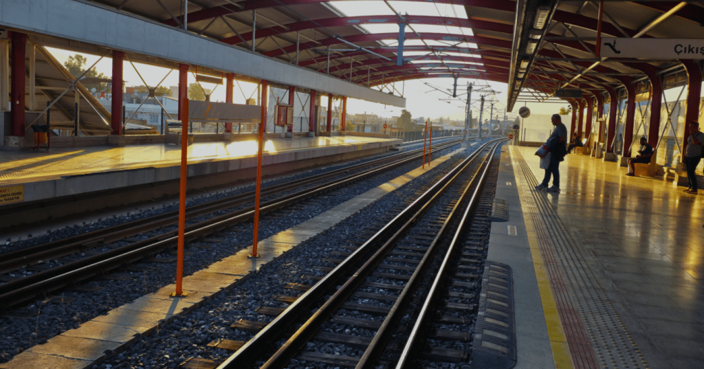 Güneş ışığında parlayan bir tren istasyonu platformu, boş raylar ve bekleyen bir yolcu.