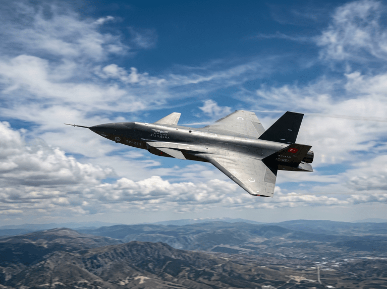 Turkish prototype stealth fighter jet flying above mountains with blue sky and clouds